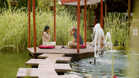 Fernansicht-Eines-Paares,-Das-Tee-Trinkt-Und-Sich-Unterhält,-Während-Sie-Auf-Kissen-Auf-Einer-Veranda-Sitzen,-Die-Von-Wasser-In-Einem-Japanischen-Garten-Umgeben-Ist