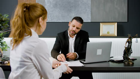 Caucasian-legal-advisor-sitting-at-a-table-with-laptop-in-the-office.-He-is-advising-a-ginger-woman