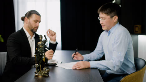 Close-up-view-of-statue-of-justice.-On-the-background-two-men-legal-advisors,-caucasian-and-asian,-talking-about-work-sitting-at-a-table-in-the-office