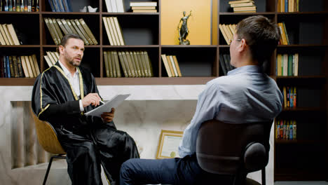 Caucasian-male-legal-advisor-wearing-toga-and-sitting-on-a-chair-in-the-office.-He-is-advising-a-client