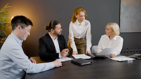 Group-of-multiethnic-legal-advisors-talking-and-reading-documents-sitting-at-a-table-in-the-office.-One-of-them-is-standing-between-two-co-workers