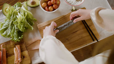 Vista-Superior-De-Las-Manos-De-Una-Mujer-Haciendo-Sushi-Sobre-Una-Tabla-De-Cocina.-En-El-Fondo,-Su-Pareja-Está-Cortando-Pescado-Fresco.