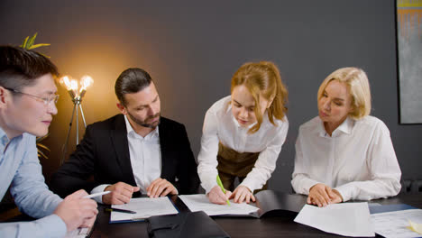 Camera-zoom-out-on-a-group-of-multiethnic-legal-advisors-talking-and-reading-documents-sitting-at-a-table-in-the-office.-One-of-them-is-standing-between-two-co-workers