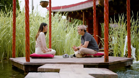 Japanese-couple-having-tea-and-talking-while-they-are-sitting-on-cushions-on-a-porch-surrounded-by-water-in-a-japanese-garden