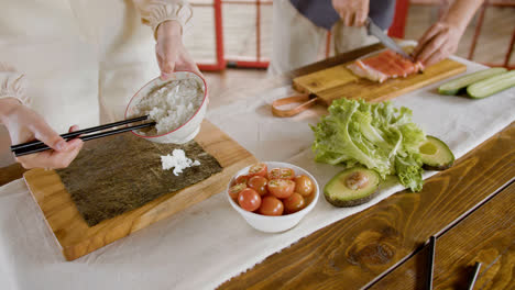 Close-up-view-of-hands-of-a-woman-making-sushi-on-top-of-a-kitchen-board.-On-the-background-her-partner-is-cutting-fresh-fish