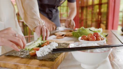 Close-up-view-of-hands-of-a-woman-making-sushi-on-top-of-a-kitchen-board.-On-the-background-her-partner-is-cutting-fresh-fish