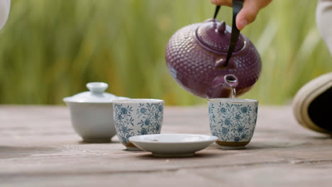 Vista-De-Cerca-De-Las-Manos-De-Un-Hombre-Japonés-Sentado-En-Un-Jardín-Japonés-Sirviendo-Té-En-Tazas.-Luego,-él-Y-Una-Mujer-Recogen-Una-Taza-De-Té-Del-Suelo.