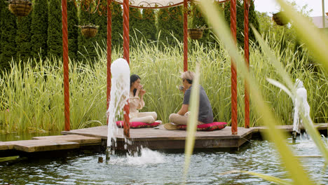 Fernansicht-Eines-Paares,-Das-Tee-Trinkt-Und-Sich-Unterhält,-Während-Sie-Auf-Kissen-Auf-Einer-Veranda-Sitzen,-Die-Von-Wasser-In-Einem-Japanischen-Garten-Umgeben-Ist