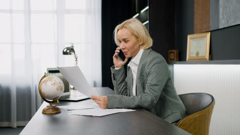 Side-view-of-caucasian-female-legal-advisor-wearing-elegant-clothes-talking-on-the-phone-and-sitting-a-table-in-the-office