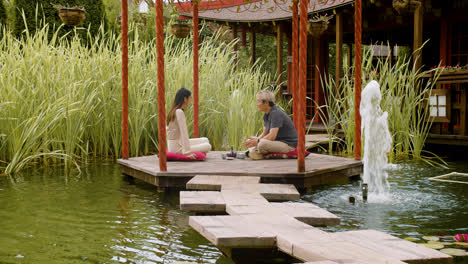 Fernansicht-Eines-Paares,-Das-Tee-Trinkt-Und-Sich-Unterhält,-Während-Sie-Auf-Kissen-Auf-Einer-Veranda-Sitzen,-Die-Von-Wasser-In-Einem-Japanischen-Garten-Umgeben-Ist