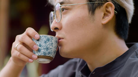 Close-up-view-of-japanese-man-in-glasses-drinking-tea-from-a-cup-while-sitting-on-a-japanese-garden-porch