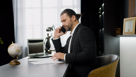 Seitenansicht-Eines-Kaukasischen-Männlichen-Rechtsberaters-In-Schwarzem-Anzug,-Der-Am-Telefon-Spricht-Und-An-Einem-Tisch-Im-Büro-Sitzt