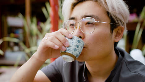 Close-up-view-of-japanese-man-in-glasses-drinking-tea-from-a-cup-while-sitting-on-a-japanese-garden-porch