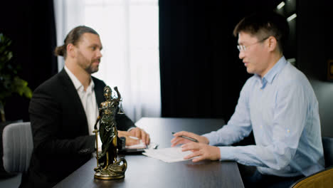 Close-up-view-of-statue-of-justice.-On-the-background-two-men-legal-advisors,-caucasian-and-asian,-talking-about-work-sitting-at-a-table-in-the-office