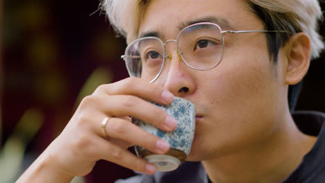 Close-up-view-of-japanese-man-in-glasses-drinking-tea-from-a-cup-while-sitting-on-a-japanese-garden-porch