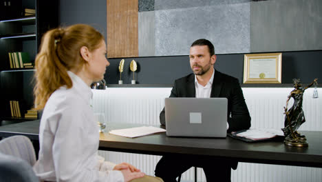 Caucasian-legal-advisor-sitting-at-a-table-with-laptop-in-the-office.-He-is-advising-a-ginger-woman