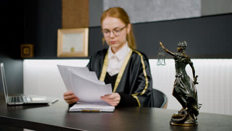 Nahaufnahme-Einer-Statue-Der-Gerechtigkeit.-Im-Hintergrund-Eine-Kaukasische-Rechtsberaterin-In-Toga,-Die-An-Einem-Tisch-Im-Büro-Sitzt-Und-Ein-Dokument-Liest.