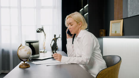 Side-view-of-caucasian-female-legal-advisor-wearing-elegant-clothes-talking-on-the-phone-and-sitting-a-table-in-the-office