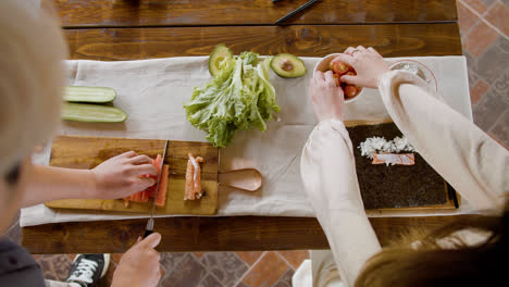 Vista-Superior-De-Las-Manos-De-Una-Mujer-Haciendo-Sushi-Sobre-Una-Tabla-De-Cocina.-En-El-Fondo,-Su-Pareja-Está-Cortando-Pescado-Fresco.