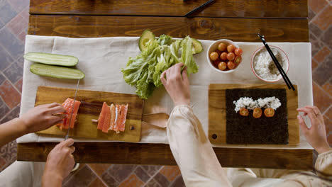 Vista-Superior-De-Las-Manos-De-Una-Mujer-Haciendo-Sushi-Sobre-Una-Tabla-De-Cocina.-En-El-Fondo,-Su-Pareja-Está-Cortando-Pescado-Fresco.