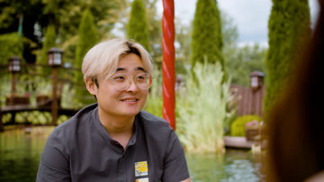 Vista-Trasera-De-Una-Mujer-Hablando-Con-Un-Hombre-Japonés-Con-Gafas-En-El-Porche-De-Un-Jardín-Japonés-Mientras-Beben-Té