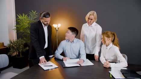 Asian-man-signing-a-document-sitting-at-table-in-the-office-while-legal-advisors-supervise