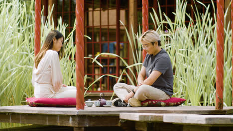 Vista-Lejana-De-Una-Pareja-Tomando-Té-Y-Conversando-Mientras-Están-Sentados-Sobre-Cojines-En-El-Porche-De-Un-Jardín-Japonés.