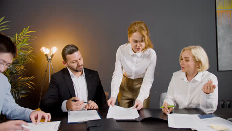 Camera-zoom-on-a-group-of-multiethnic-legal-advisors-talking-and-reading-documents-sitting-at-a-table-in-the-office.-One-of-them-is-standing-between-two-co-workers