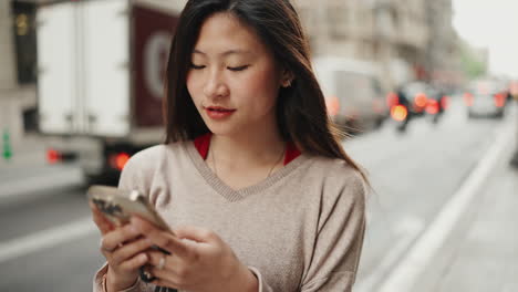 Mujer-Asiática-Joven-Sonriendo-Mientras-Usa-Un-Teléfono-Inteligente-Al-Aire-Libre.