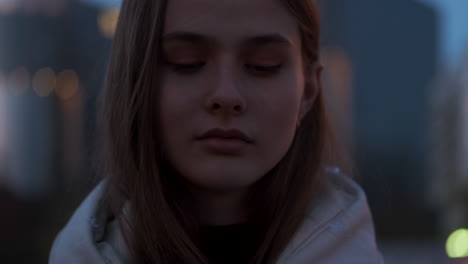 Portrait-of-confident-Caucasian-girl-intently-looking-in-camera-on-evening-street-at-dusk-city.