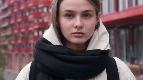 Portrait-of-serious-Caucasian-girl-in-a-jacket-with-scarf-confidently-looking-in-camera-on-city-street.