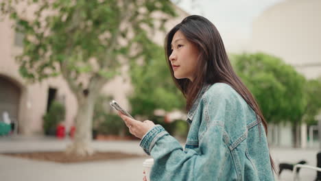 Asiatische-Frau-Checkt-Ihr-Telefon,-Während-Sie-Draußen-Sitzt.