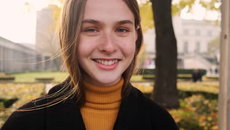 Retrato-De-Una-Alegre-Niña-Caucásica-Mirando-Felizmente-A-La-Cámara-Y-Sonriendo-En-El-Parque-De-La-Ciudad-Caminando-Al-Atardecer.