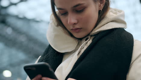 Close-up-shot-of-casual-girl-in-down-jacket-with-scarf-intently-using-cellphone-at-metro-station.