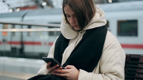 Medium-shot-of-Caucasian-girl-in-a-jacket-with-scarf-thoughtfully-using-cellphone-waiting-at-subway-station.