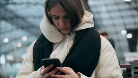 Chica-Caucásica-Con-Chaqueta-De-Plumas-Y-Bufanda-Usando-Un-Teléfono-Celular-Esperando-El-Tren-En-La-Estación-De-Metro.