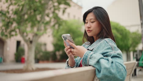 Asiatische-Frau-Checkt-Ihr-Telefon,-Während-Sie-Auf-Einer-Bank-Im-Park-Sitzt.