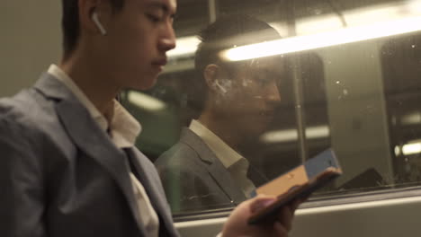 Joven-Empresario-Asiático-Confiado-Con-Auriculares-Inalámbricos-Usando-Un-Teléfono-Celular-En-El-Tren-Subterráneo.