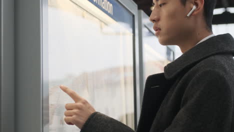 Young-Asian-businessman-in-coat-with-wireless-earphones-watching-public-transport-route-waiting-on-street.