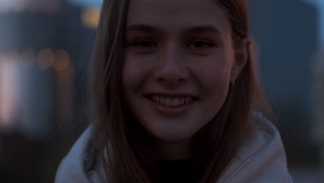 Close-up-shot-of-Caucasian-girl-smiling-and-joyfully-looking-in-camera-on-evening-at-dusk-street.