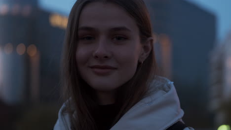 Close-up-portrait-shot-of-Caucasian-girl-happily-smiling-in-camera-on-evening-street-walking-at-dusk.