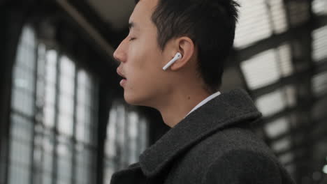 Young-Asian-man-in-coat-using-wireless-earphones-waiting-train-at-subway-station.