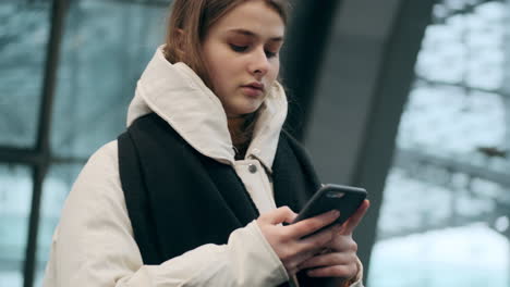 Kaukasisches-Mädchen-In-Daunenjacke-Und-Schal-Benutzt-Ein-Mobiltelefon-Und-Schaut-Nachdenklich-Zur-Seite,-Während-An-Der-U-Bahn-Station-Ein-Wartender-Zug-Wartet.