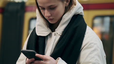 Serious-Caucasian-girl-in-down-jacket-with-scarf-intently-using-cellphone-sitting-at-subway-station.