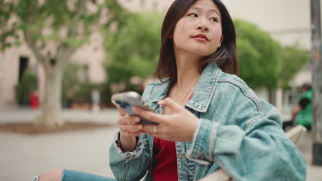 Asiatische-Frau-Schaut-Sich-Um-Und-Checkt-Ihr-Telefon,-Während-Sie-Draußen-Sitzt.