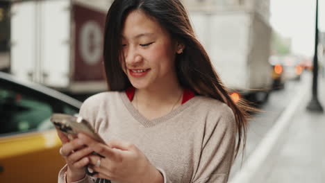 Mujer-Asiática-Joven-Sonriendo-Mientras-Usa-Un-Teléfono-Inteligente-Al-Aire-Libre.