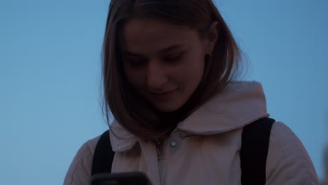 Chica-Caucásica-Felizmente-Usando-Un-Teléfono-Celular,-Sonriendo,-Parada-Sola-Al-Aire-Libre-En-La-Ciudad-Al-Anochecer.