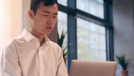 Joven-Asiático-Con-Camisa-En-Un-Café-Trabajando-Pensativamente-En-Una-Computadora-Portátil-Y-Sonriendo.