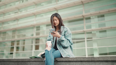 Asian-woman-drinking-coffee-and-checking-her-phone-outdoors.