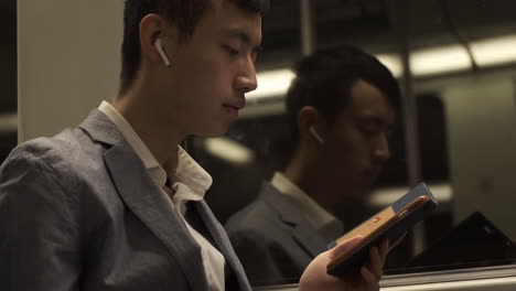 Young-Asian-businessman-in-wireless-earphones-thoughtfully-using-cellphone-in-subway-train.